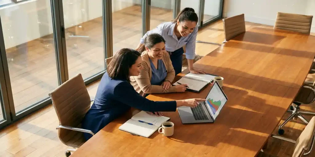 Drie verpleegkundigen bespreken werk rond laptop aan vergadertafel met notitieblokken en koffie in licht kantoor