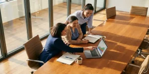 Drie verpleegkundigen bespreken werk rond laptop aan vergadertafel met notitieblokken en koffie in licht kantoor