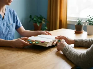 Zorgverlener houdt opengeslagen boek met kleurrijke bladwijzers vast terwijl patiënt handen uitstrekt over houten consultatietafel